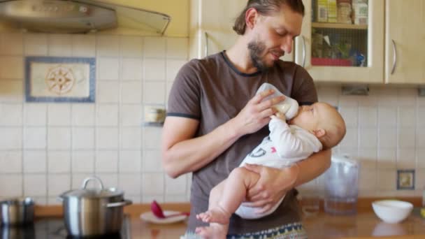 De vader van een jonge multitasking en zuigeling in de keuken. De man eindigt met het voeden van de baby, neemt de fles en beide glimlach. Man en kind in een moderne keuken — Stockvideo