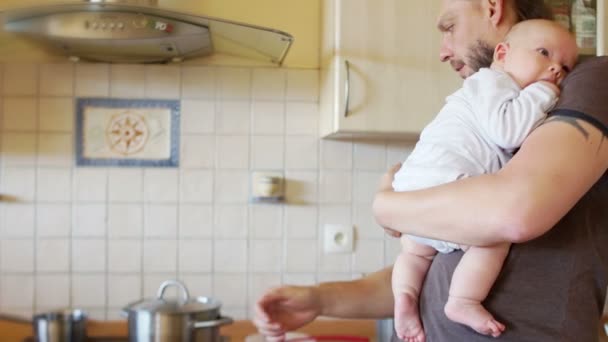 Un hermoso bebé en los brazos de un padre joven. Un hombre multitarea está alimentando al bebé en la cocina y cocinando simultáneamente sopa. Día del Padre — Vídeo de stock