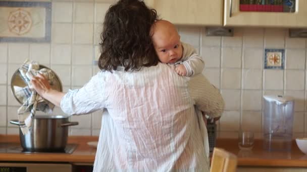 Joven ama de casa preparando la cena en la cocina. Una mujer sostiene a un bebé en sus manos. Multitarea: alimenta al bebé y cocina la sopa en la estufa. Día de las Madres — Vídeos de Stock