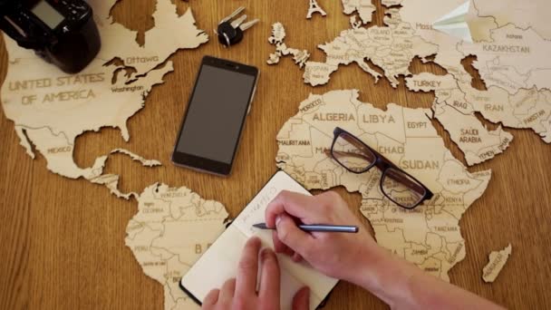 A close portrait of a man writes a plan for his journey. Wooden world map layout on the table, notebook, pen, phone, keys, glasses and camera — Stock Video