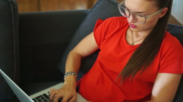 La morena vestida de rojo está escribiendo texto en el teclado del portátil, sentada en una silla. Trabajo a distancia, una mujer freelancer, redes sociales — Vídeos de Stock