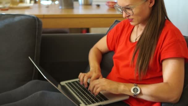 Una ragazza in abito rosso è in corrispondenza con i social network, seduta comodamente su una sedia morbida. Donna freelance. Lavoro a distanza, lavoro a distanza, lavoro tramite laptop — Video Stock