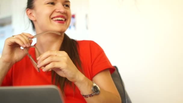 Jonge heldere brunette in het kantoor. Het meisje is gekleed in een rode jurk. Breken in werk, kijkt weg en lacht, kleren inzetglazen — Stockvideo