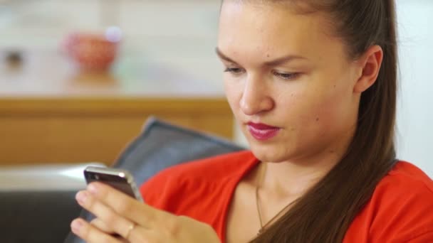 Una joven con un vestido rojo sonríe en la pantalla de un teléfono móvil. Levanta la cabeza y sonríe. Redes sociales, comunicación en redes sociales. Primer plano, movimiento descuidado — Vídeos de Stock