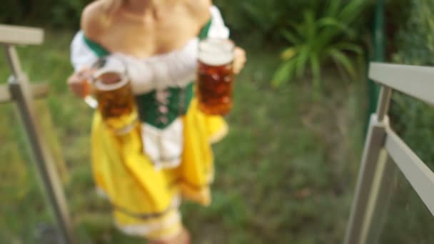 Сексуальная Октоберфест официантка несет два больших бокала пива. Девушка ходит босиком по траве и поднимается по лестнице. Баварский традиционный костюм, эротический вырез — стоковое видео