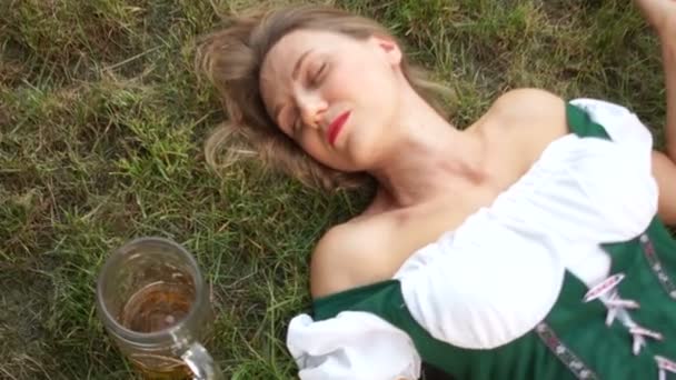 A lovely waitress in a national Bavarian dress at the Oktoberfest festival has drunk too much beer and is sleeping on the grass — Stock Video