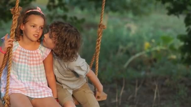 Un chico y una chica cariñosos montan en un columpio sentados uno al lado del otro. El chico besa a la chica, ella lo golpea en la mejilla, una bofetada en la cara — Vídeos de Stock