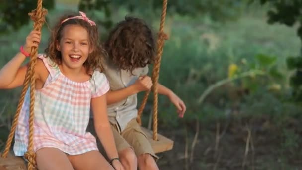 Hermano y hermana montan en un columpio. Vacaciones de verano, descanso en el pueblo, hermosos niños rizados — Vídeos de Stock