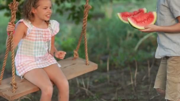 Chica feliz y niño comiendo una sandía sentado en un columpio, foto de verano. Niños con sandía — Vídeo de stock