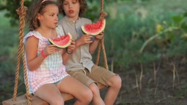 Los niños comen sandía, balanceándose en un columpio. Cálidas vacaciones de verano, familia feliz, hermano y hermana — Vídeo de stock