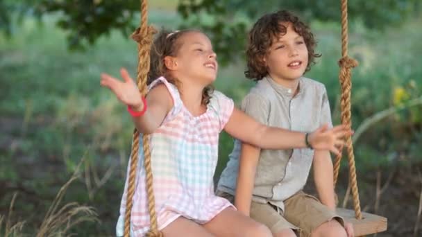 Dziewczyna i chłopak, dzieci w wieku szkolnym, brat i siostra mówimy entuzjastycznie, siedząc na huśtawce w parku. Dziewczyna jest gestykulując. Letnie wakacje na świeżym powietrzu na wsi — Wideo stockowe