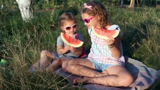 Zwei Schwestern sonnen sich in der Sonne und essen eine Wassermelone. der jüngere streckt den Anteil der Älteren aus und nimmt ihn an sich. der Älteste in Ratlosigkeit. Kinderwitz, Sommerferien — Stockvideo