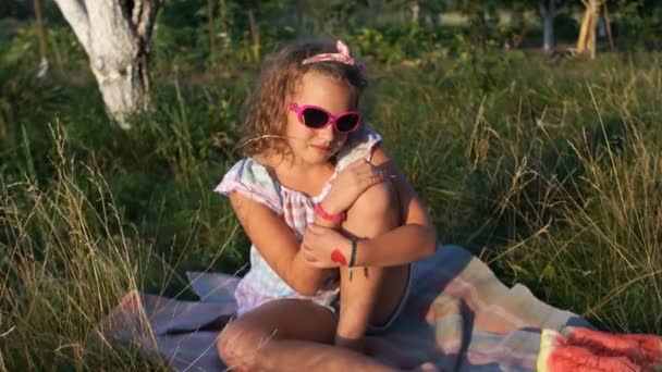 Egy iskolás, napozó ül a fűben. A lány visel, napszemüveget és a kötést a fejét. Piknik a természetben. Nyári vakáció — Stock videók