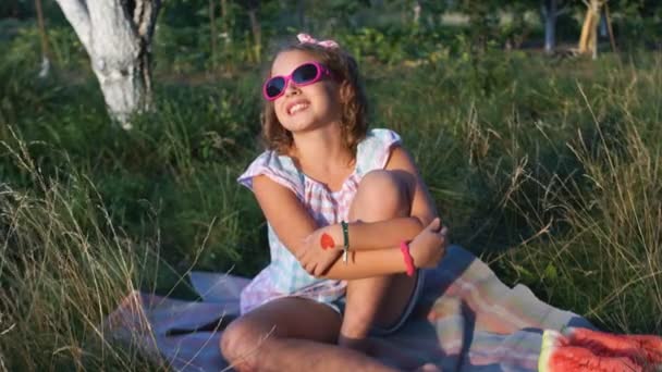 Piknik doğada. Bir genç kız köyün çimlerde oturan sunbathes. O pembe güneş gözlüğü, bir bandaj başını ve bir bilezik onun koluna takıyor — Stok video