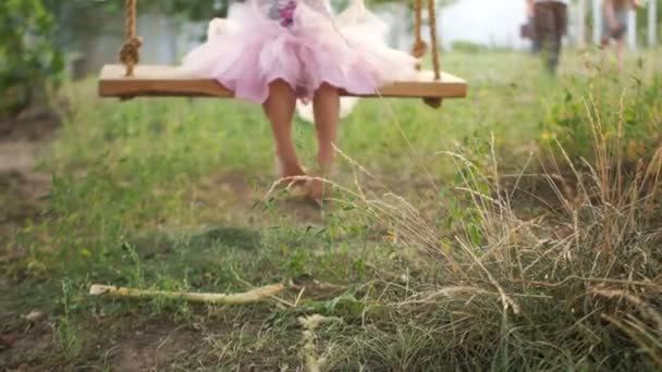 Uma menina de saia rosa tutti patina em um balanço caseiro rural. Close-up de pés descalços, grama e terra. Boas festas. — Vídeo de Stock