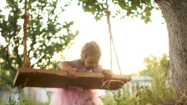 Three-year-old little girl rolls on a makeshift swing lying on her belly. A child laughs cheerfully, shows a tongue, teases — Stock Video