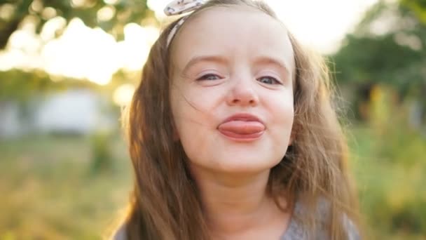 En provocerande liten fyra-åring flicka visar sin tunga. Närbild porträtt mot en sommar solnedgång bakgrund. Friska barndom — Stockvideo