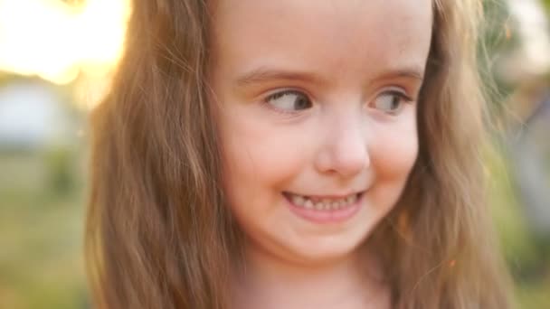 Ett nära porträtt av en liten käck långhåriga flicka. Barnet skrattar, visar de mjölka tänderna. Sommardag, solnedgång. Friska barn — Stockvideo