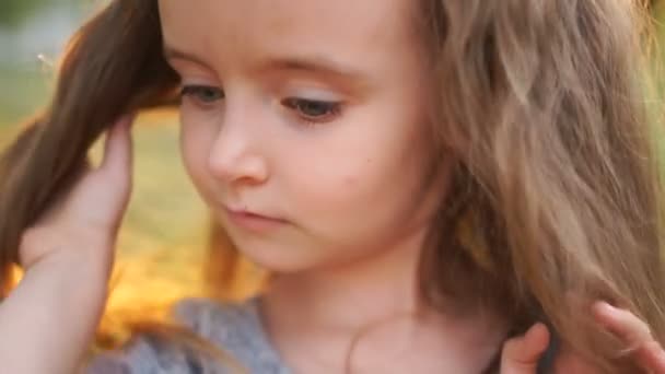 Maravilloso retrato de una hermosa niña con el pelo largo y los ojos grises. Contra el telón de fondo de una puesta de sol de verano. La niña se ajusta el pelo y baja las pestañas largas — Vídeo de stock