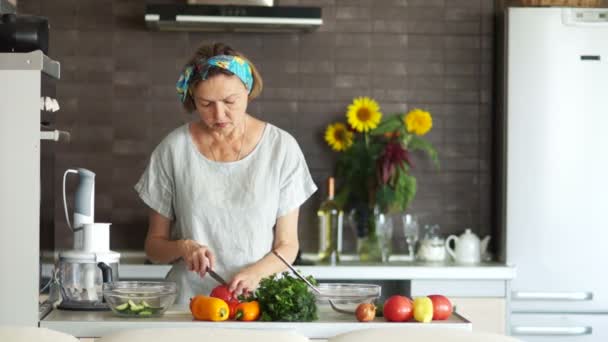 Современный пенсионер готовит салат дома на кухне. Она концентрирует помидоры. Её волосы завязаны бинтом. Правильное питание, здоровый образ жизни, рецепт приготовления салата — стоковое видео