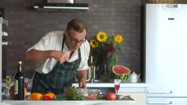 Um idoso de óculos prepara o jantar na cozinha. Mexa os legumes em uma tigela. Um pensionista com uma cara séria na cozinha, temperando uma salada. Dia das Mães — Vídeo de Stock