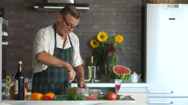 Hombre maduro en vasos prepara una ensalada en la cocina. Revuelve las verduras en un tazón. Retirado con una cara seria en la cocina, ensalada de condimentos — Vídeos de Stock