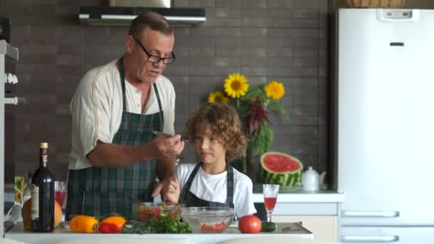 Abuelo y nieto en la cocina. Un anciano le da al niño un cuchillo de cocina y te enseña a cortar un pepino. Clase magistral del cocinero. Los hombres están cocinando. — Vídeo de stock