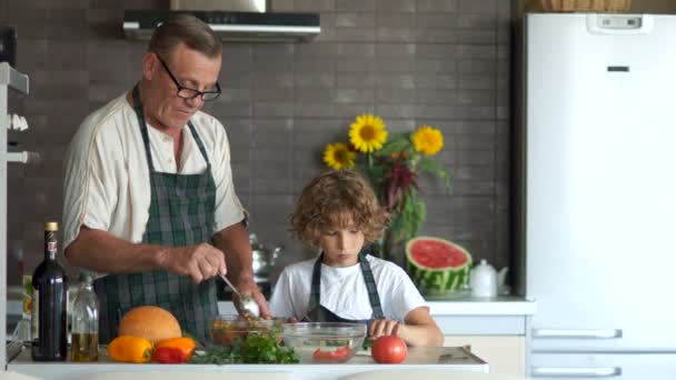 De kleinzoon snijdt een komkommer voor salade op het bord van de keuken. Grootvader vertelt hem iets. Culinaire onderwijsvaardigheden. De continuïteit van generaties — Stockvideo