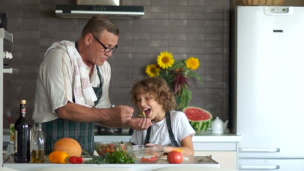 Дедушка и внук вместе готовят ужин. Мальчик режет сладкий перец для салата. Мужчина дает салат мальчику. Мастер-класс — стоковое видео