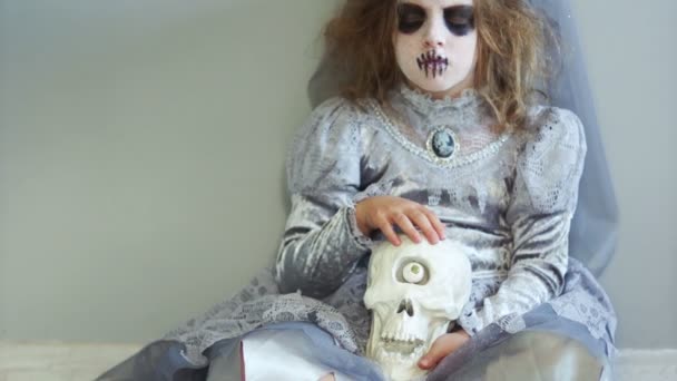 Colegiala con un traje de bruja gris con una corona en la cabeza se sienta y sostiene un cráneo con un ojo en su regazo. Poco convencional halloween creativo, el día de los muertos — Vídeos de Stock