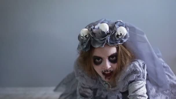 Halloween holiday. A dead bride crawls across the floor directly to the camera. A girl dressed as a dracula bride. Gray dress and veil. Wreath with skulls — Stock Video