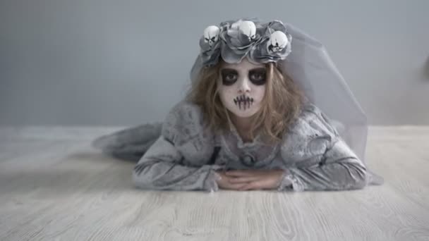 A dead bride crawls across the floor directly to the camera. A girl dressed as a dracula bride. Gray dress and veil. Wreath with skulls. Halloween holiday — Stock Video