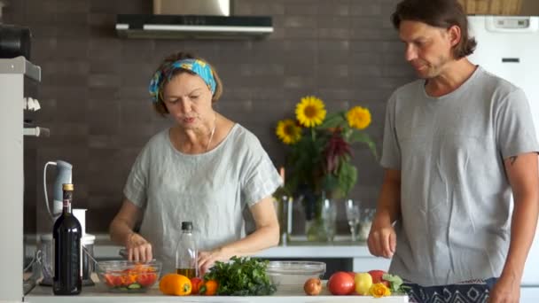 Die Rentnerin schneidet in der Küche einen Salat und tanzt lustig. Neben ihr tanzt ihr erwachsener Sohn. Altersunterschied — Stockvideo