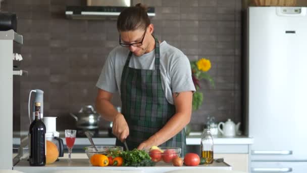 Giovane uomo serio con gli occhiali e un grembiule sta tagliando un'insalata mentre in piedi nella sua cucina. Lo chef balla con la musica. Festa dei padri, l'uomo in cucina — Video Stock