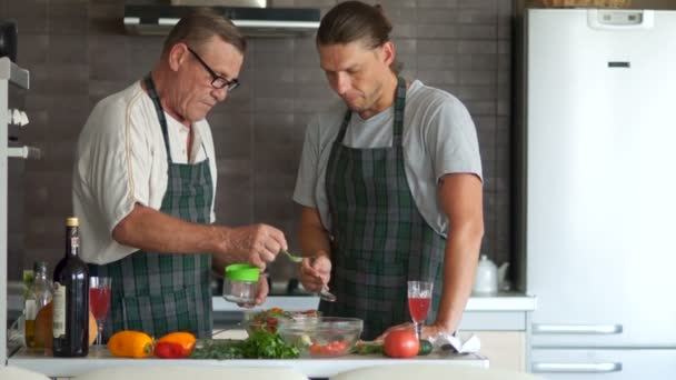 Un padre anziano con un figlio adulto sta preparando un'insalata vegetale vegetariana. Padre sale, figlio annuisce. Uomini in cucina — Video Stock