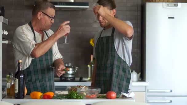 Mannen koken diner samen. Een bejaarde gepensioneerde m/v en een jonge man een plantaardige salade in de keuken te proeven. Beide lach vrolijk. Gelukkig, vaders familiedag, thanksgiving day — Stockvideo