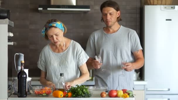 Un giovanotto e una donna matura stanno cucinando la cena in cucina. Mette gli occhiali sul tavolo. Sul tavolo ci sono verdure mature e una bottiglia di vino. Coppia con una differenza di età — Video Stock