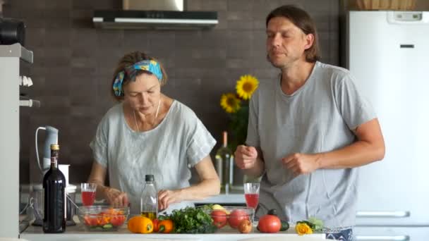 Una pareja con una diferencia de edad preparar la ensalada en la cocina, se relajan con la música y beber vino de las copas — Vídeo de stock