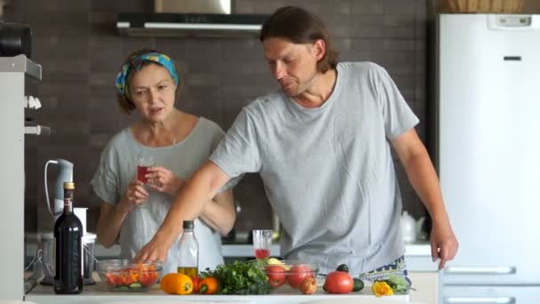Um menino e uma mulher madura estão cozinhando o jantar em uma cozinha moderna. Ele ri e brinca dançando com uma faca. Bom humor, senso de humor — Vídeo de Stock