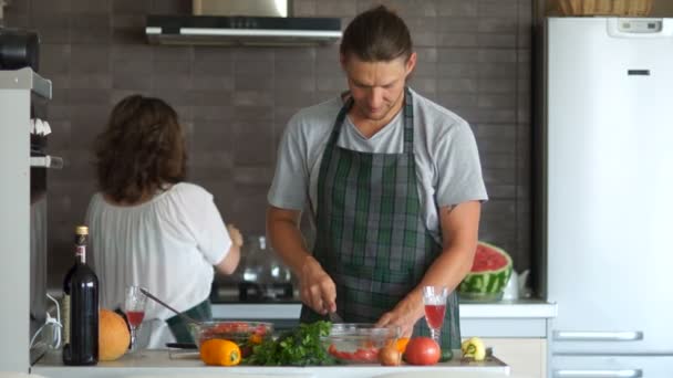 Ele corta a salada, ela cozinha a sopa no fogão na panela. Marido e mulher na cozinha preparando o jantar. A rapariga traz a panela, retira a tampa, o tipo atira um pedaço de pimenta para lá. Humor familiar — Vídeo de Stock