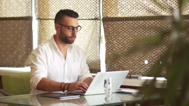 Joven barbudo trabajando en la oficina, portátil, mirando cuidadosamente a través de gafas, sonriendo. Freelancer, persianas romanas — Vídeo de stock