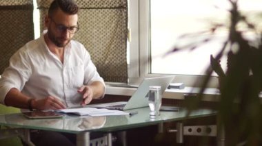 Deneyimli serbest meslek coworking içinde çalışır. Bir erkek, beyaz bir gömlek ve gözlük bir dizüstü bilgisayar yakınındaki bir masada oturan evrak gider. Belge akışı, muhasebe belgelerinin
