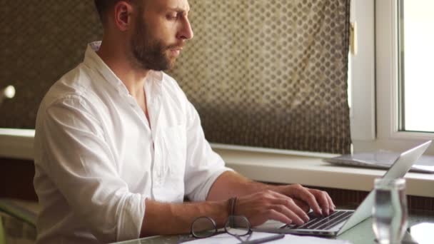 Volné noze sedící u stolu s přenosným počítačem. Mladý muž v bílé košili a stylové vousy klade na brýle na zrak — Stock video