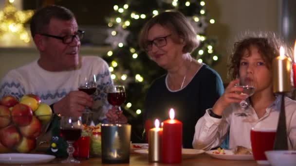 Μεγάλη οικογένεια που συγκεντρώθηκαν στο χριστουγεννιάτικο τραπέζι, ένα οικογενειακό δείπνο. Γιαγιά και ο παππούς με τον εγγονό στο τραπέζι. Η γυναίκα είναι το γέλιο. Ενήλικες πίνουν κόκκινο κρασί από ποτήρια, ποτά νερό αγόρι — Αρχείο Βίντεο