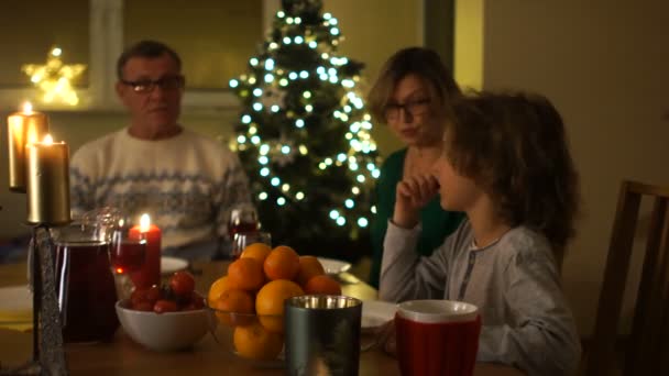 Una familia encantadora sentada a la mesa. Las luces en la habitación están apagadas, las velas en la mesa y las luces del árbol de Navidad están ardiendo. Chico se regocija en un nuevo plato sobre la mesa — Vídeos de Stock