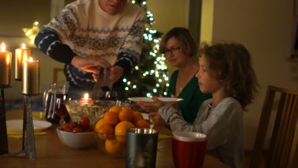 Rodzina razem Boże Narodzenie celebracja koncepcja. Dziadek stawia Sałatka na płytce wnuków. Na stole są oświetlone świecami i mandarynki są w wazonie — Wideo stockowe