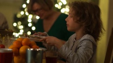 İle onun büyük bir yılbaşı ağacı arka plan üzerinde Noel masada okul çocuğu. Çocuk onun plaka yiyecek yeni bir kısmı için dışarı tutar