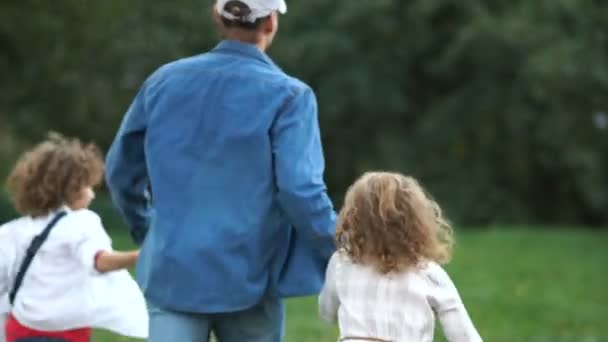 Un niño, una niña y un hombre con un perro con una correa corriendo a través de la hierba verde, vista trasera. Día de los padres familia feliz — Vídeo de stock