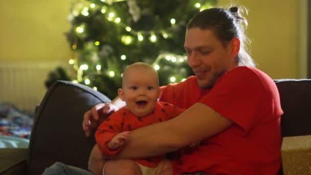 Padre y bebé están sentados en el sofá cerca del árbol de Navidad. Familia feliz, el bebé de seis meses se ríe, primera Navidad — Vídeos de Stock