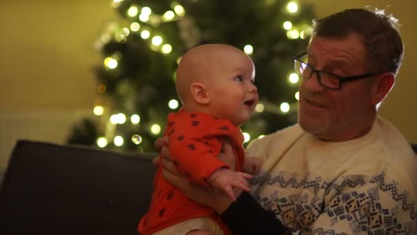 Avô beija neto, ele está sentado em um sofá contra o fundo das luzes da árvore de Natal. Duas gerações, bebé e avô — Vídeo de Stock